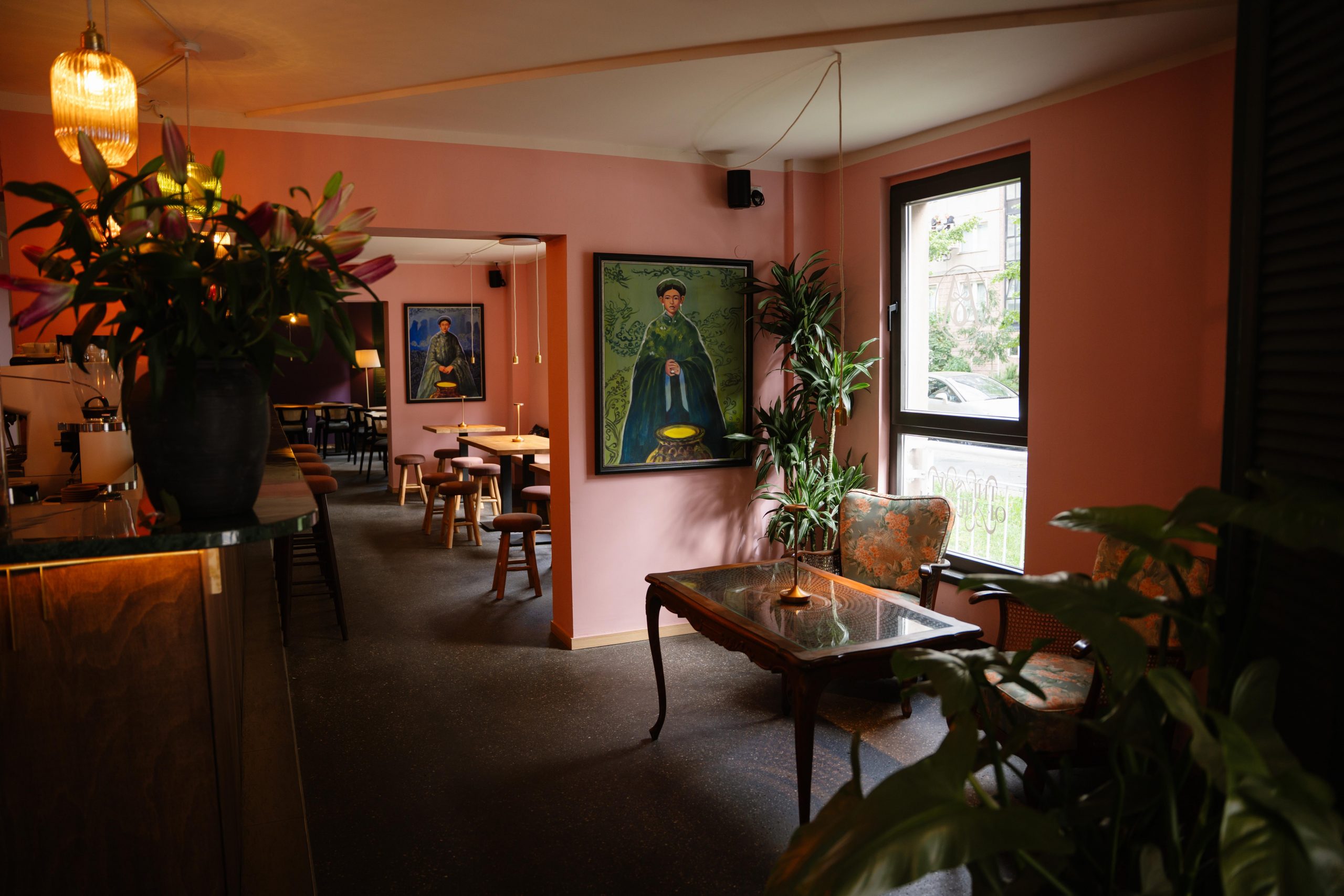 Cozy cafe with pink walls, framed portraits, plants, and a glass-top table near a sunlit window.