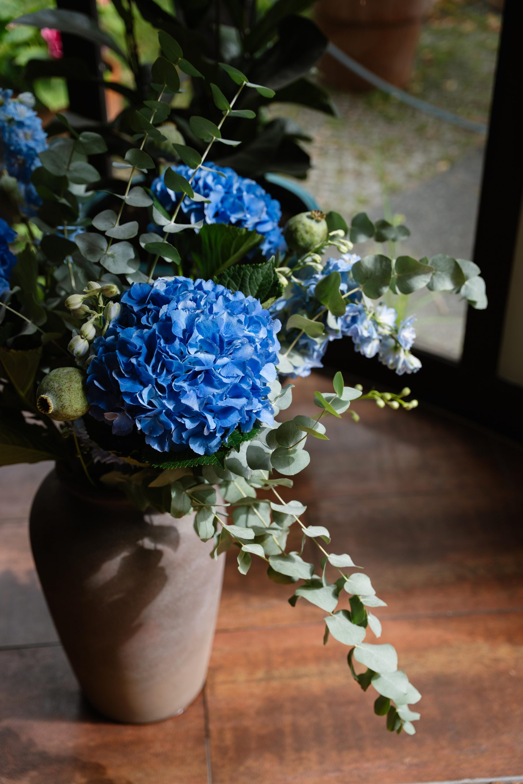 Blue hydrangea bouquet with eucalyptus in a gray vase on a wooden floor by a window.
