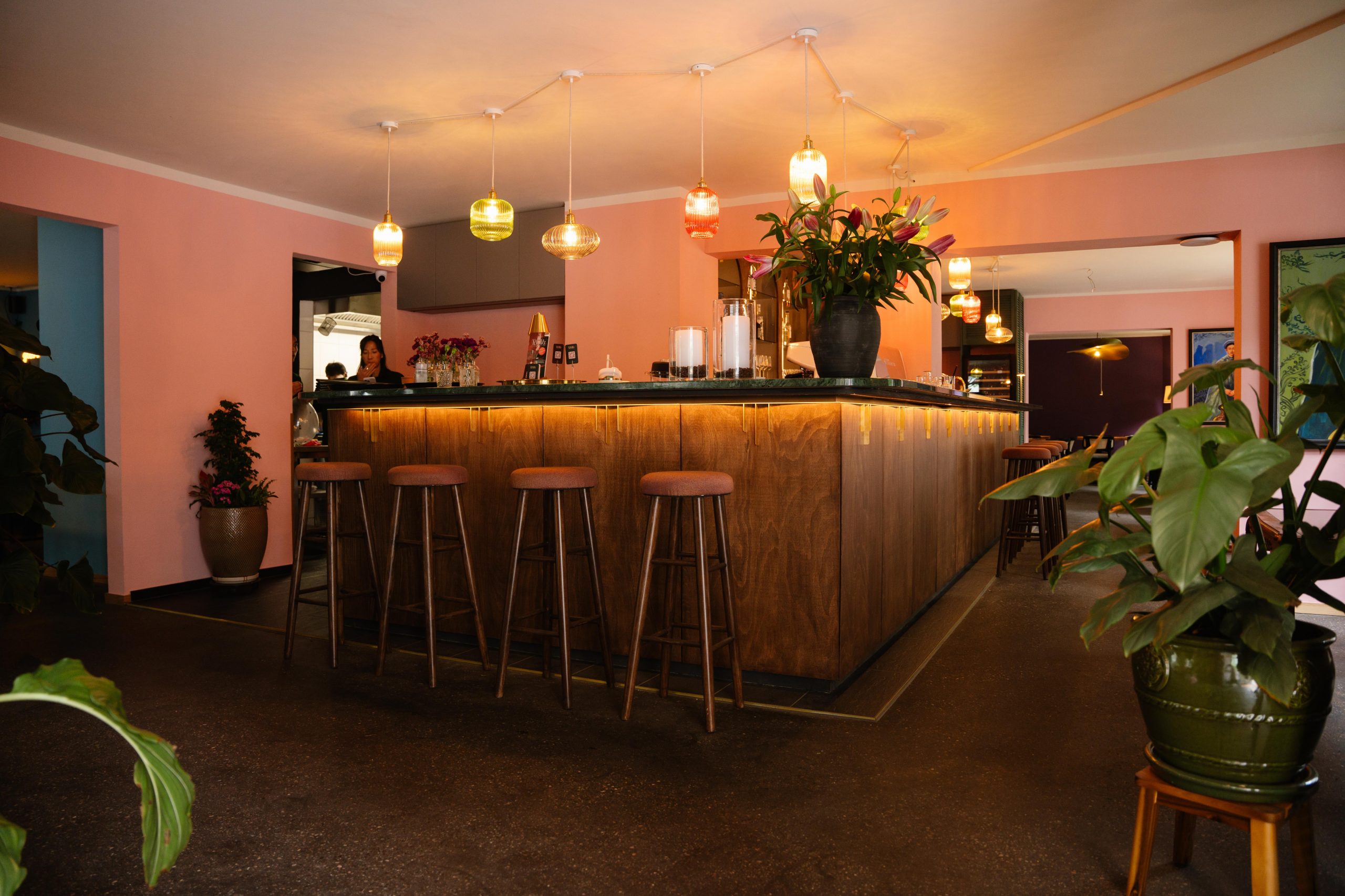 Cozy bar interior with pink walls, a long wooden counter, and multi-colored pendant lights above.