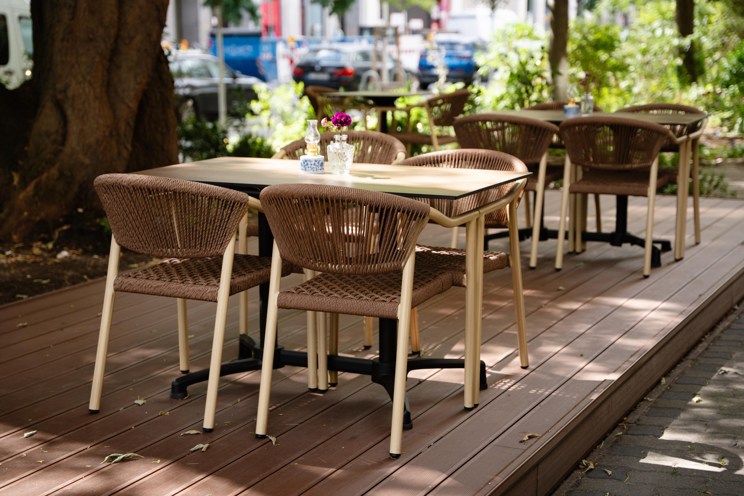 Outdoor cafe seating on a wooden deck with brown wicker chairs and light-colored tables under trees, a small vase of flowers on a table nearby.