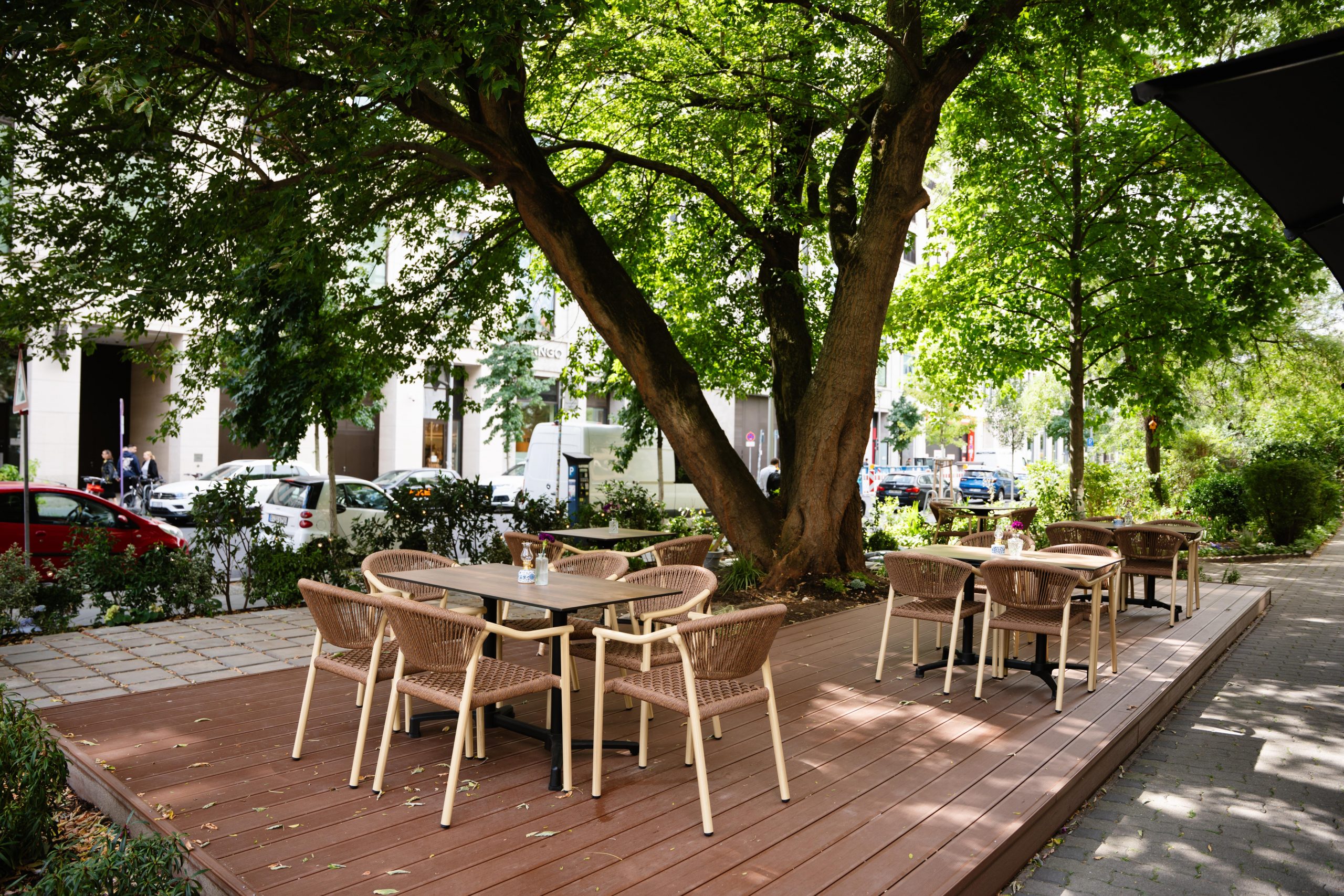 Outdoor cafe seating on a raised wooden deck under a large tree; wicker chairs surround several tables, with a city street in the background.