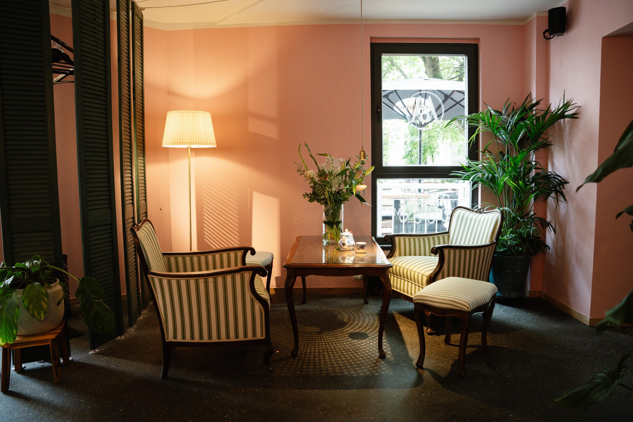 Cozy sitting area with pink walls, striped armchairs, a wooden coffee table, and a vase of flowers by a large window.