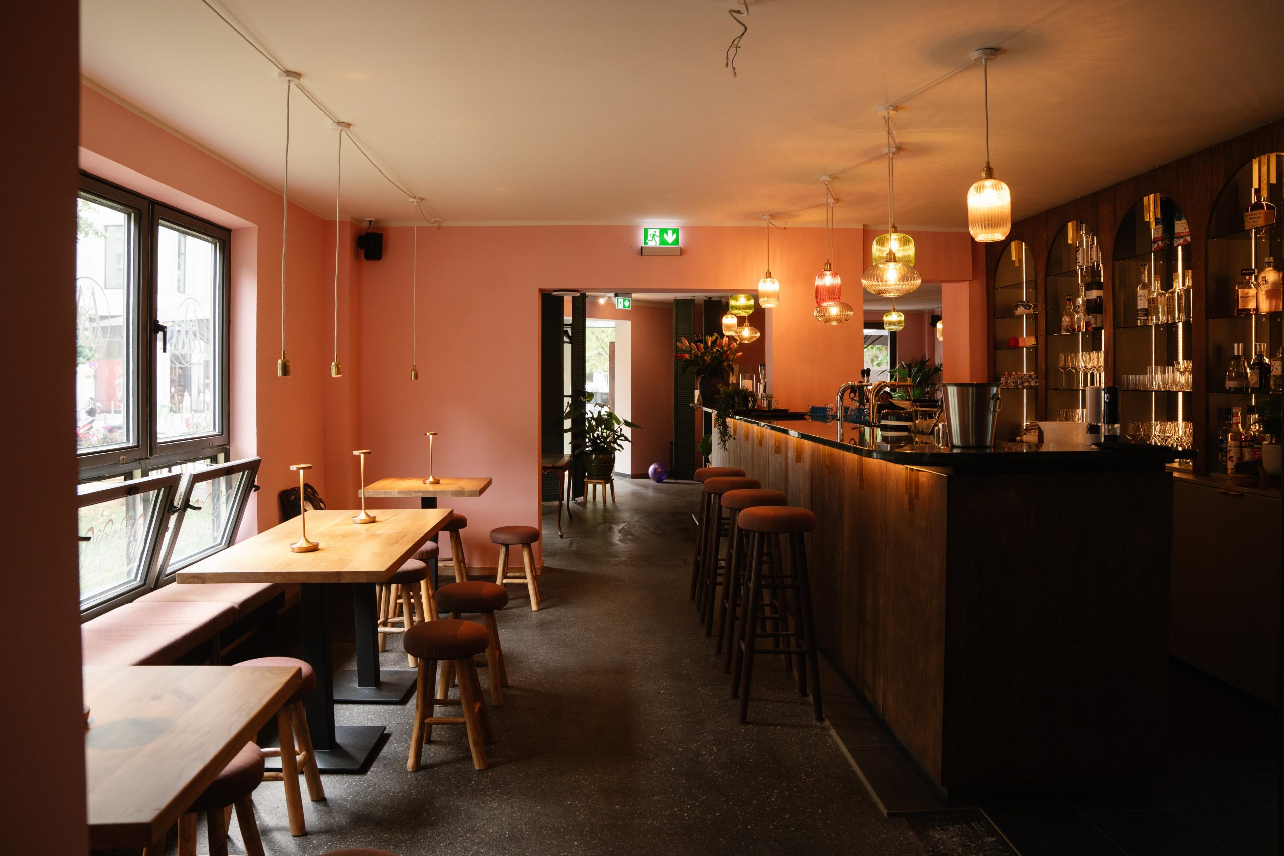 Cozy cafe interior with pink walls, wooden tables and stools along a windowed wall, and a long bar with glassware on the right.
