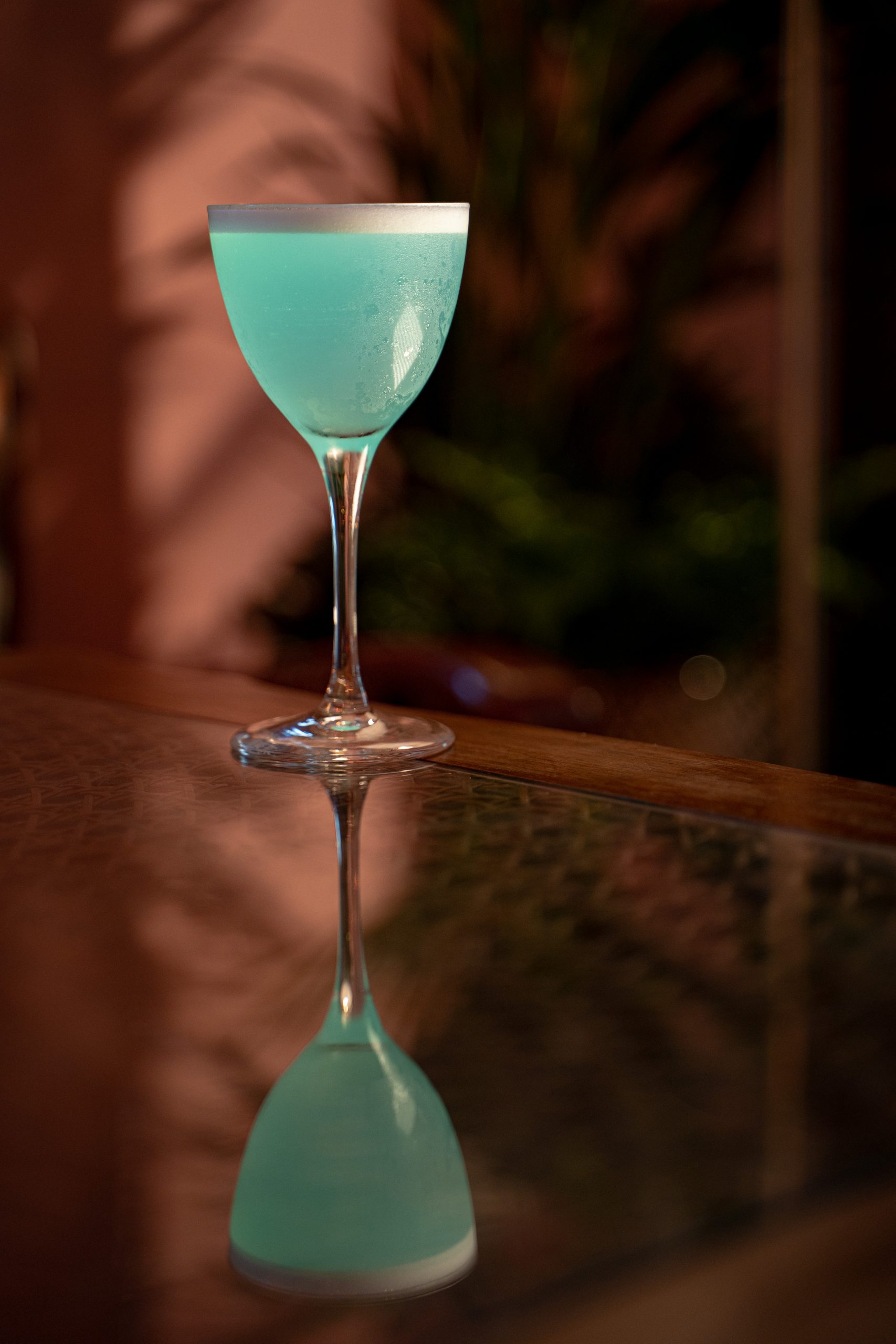 Turquoise cocktail in a stemmed coupe glass on a glossy bar, reflected below in a dimly lit setting.