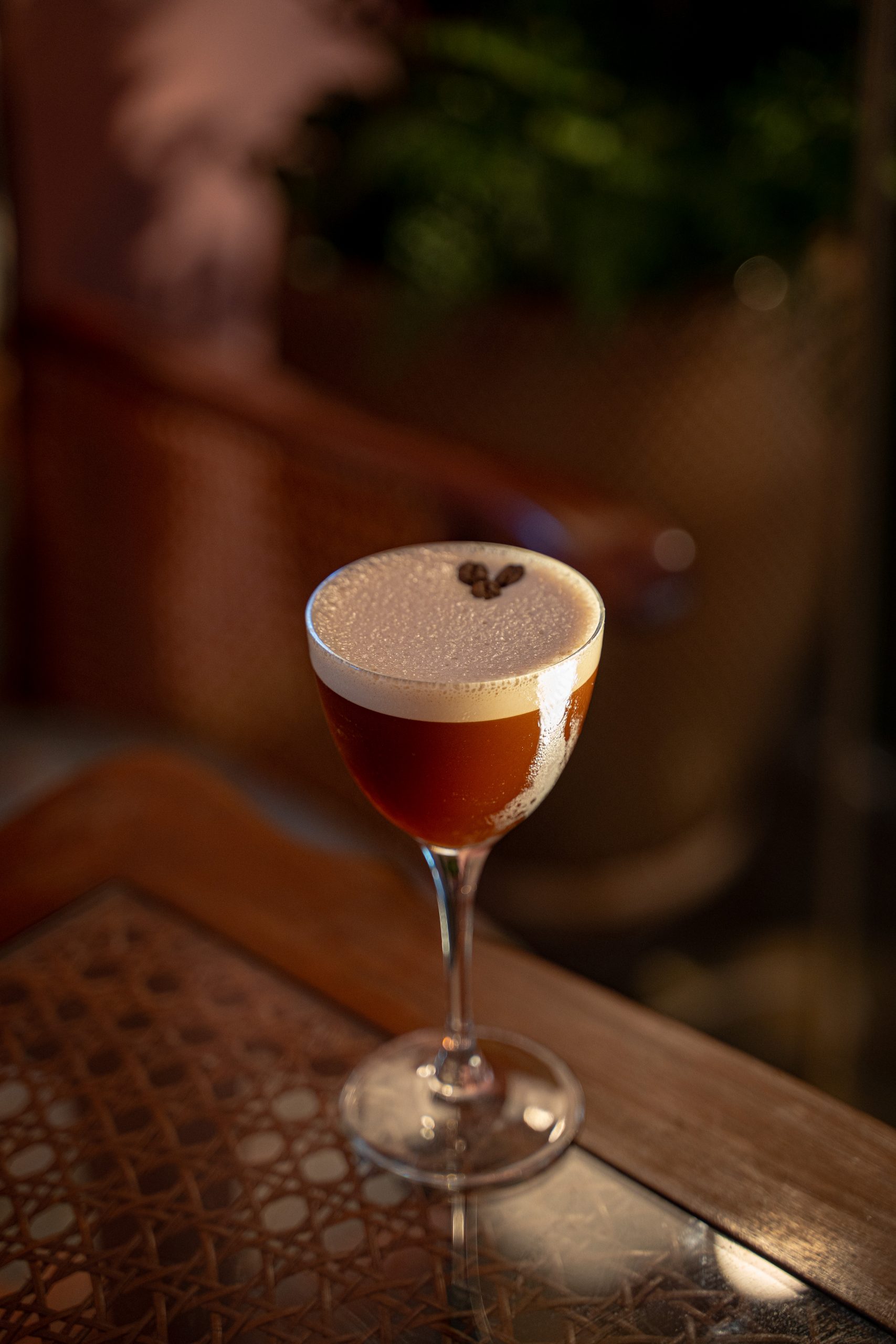 Amber cocktail in a stemmed glass with a foamy white head topped by two coffee beans, on a wooden table at night.