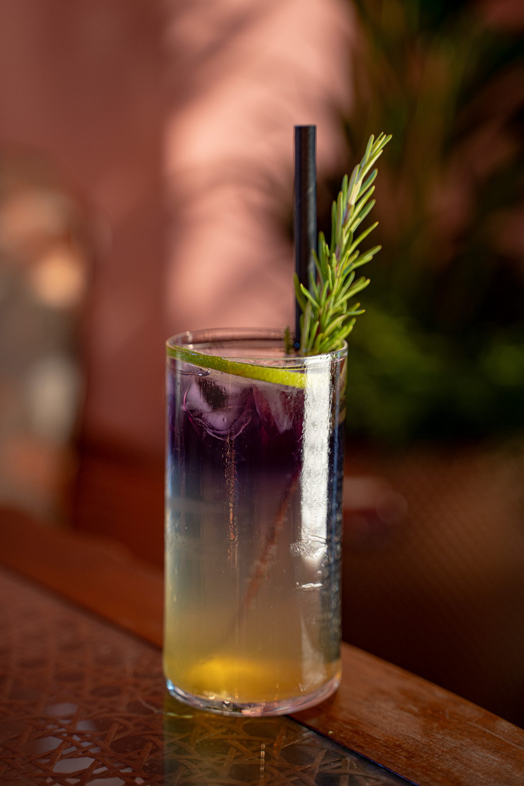 Tall glass cocktail with yellow-to-purple gradient, ice, lime wedge, rosemary garnish, and a black straw on a wooden bar surface.