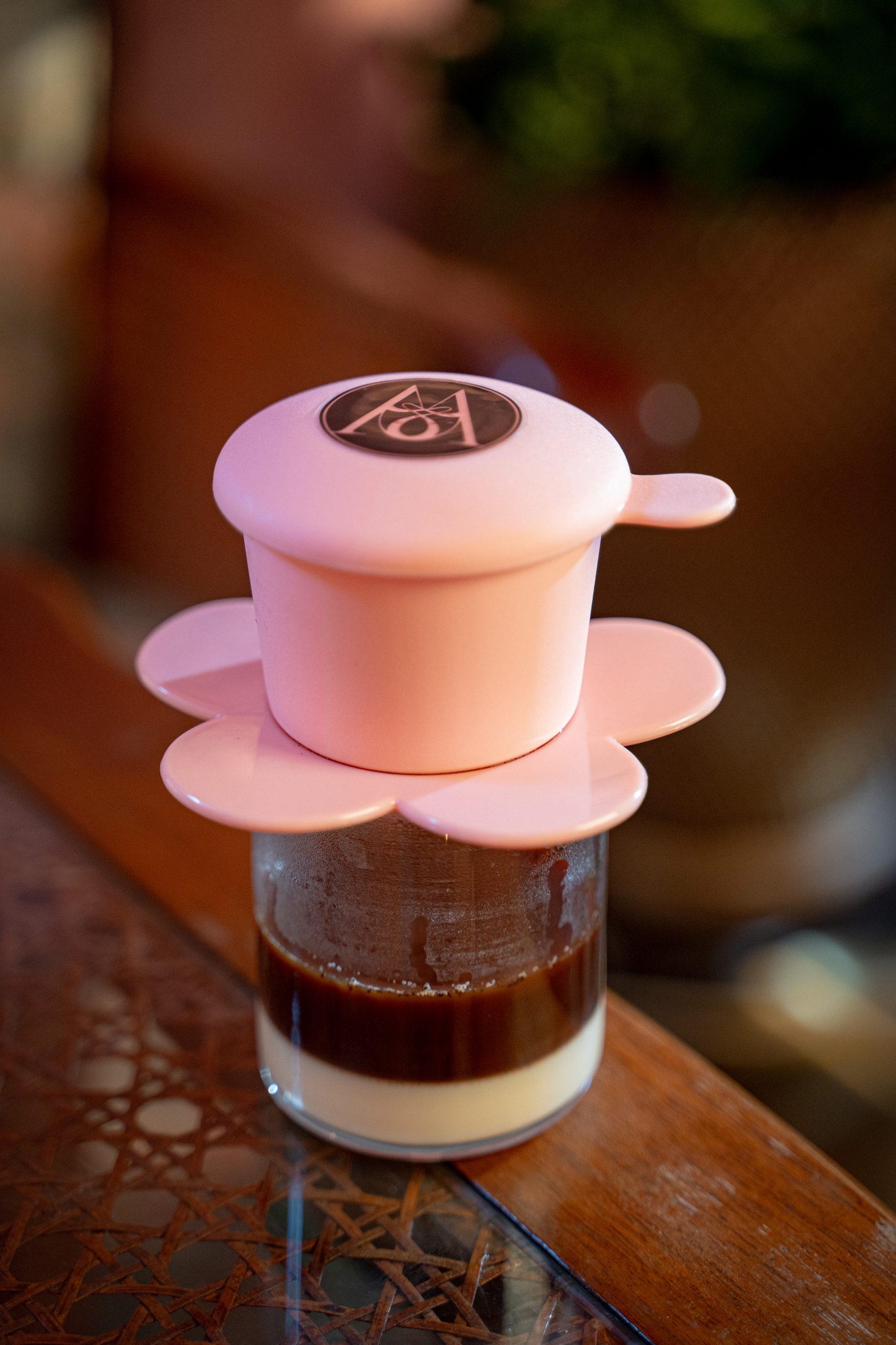Layered coffee drink in a small glass with a pink flower-shaped lid featuring a logo on top, placed on a wooden table.