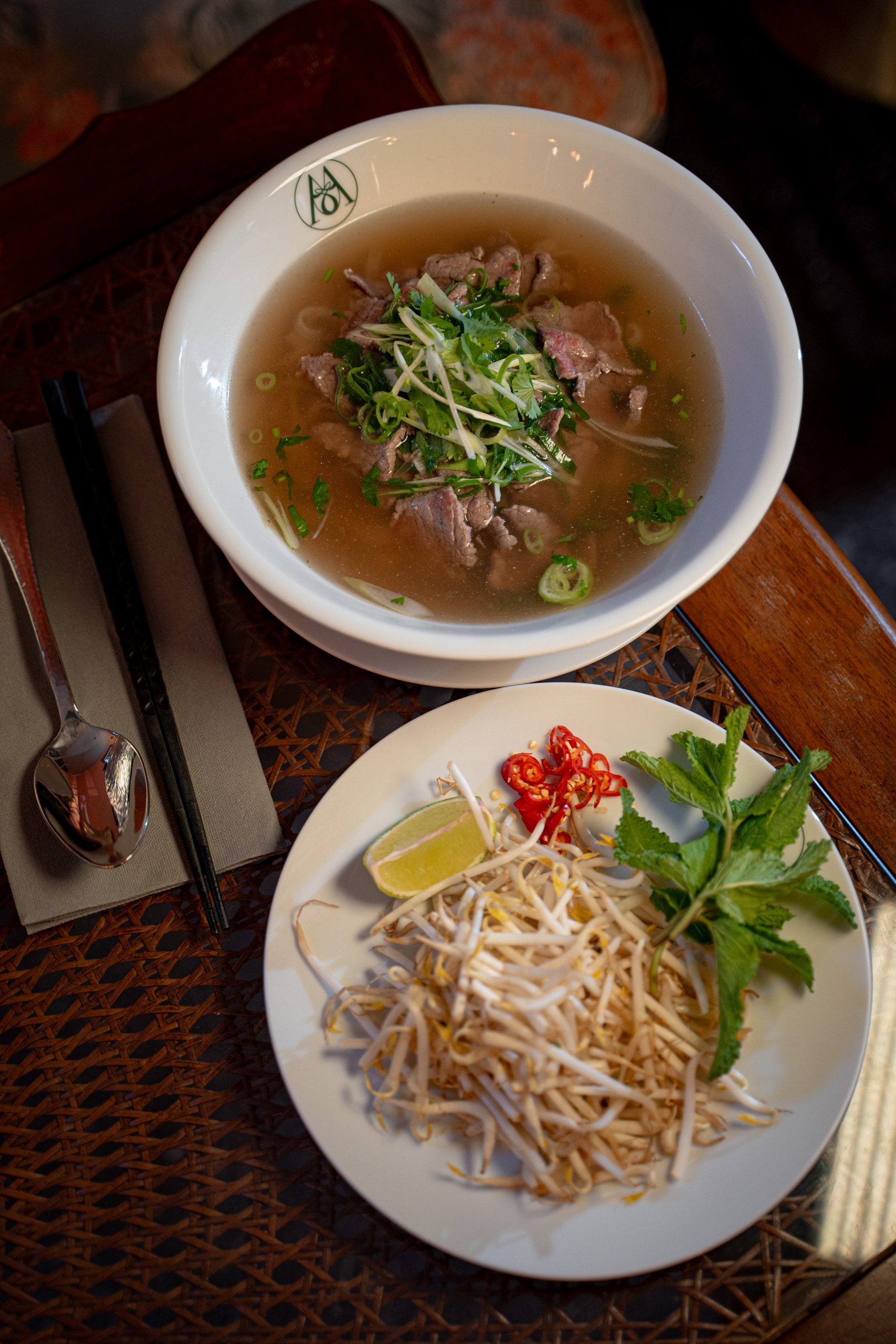 Beef pho soup with sliced beef, green onions, and cilantro, served with a side plate of bean sprouts, lime, mint, and red chilies on a white plate.
