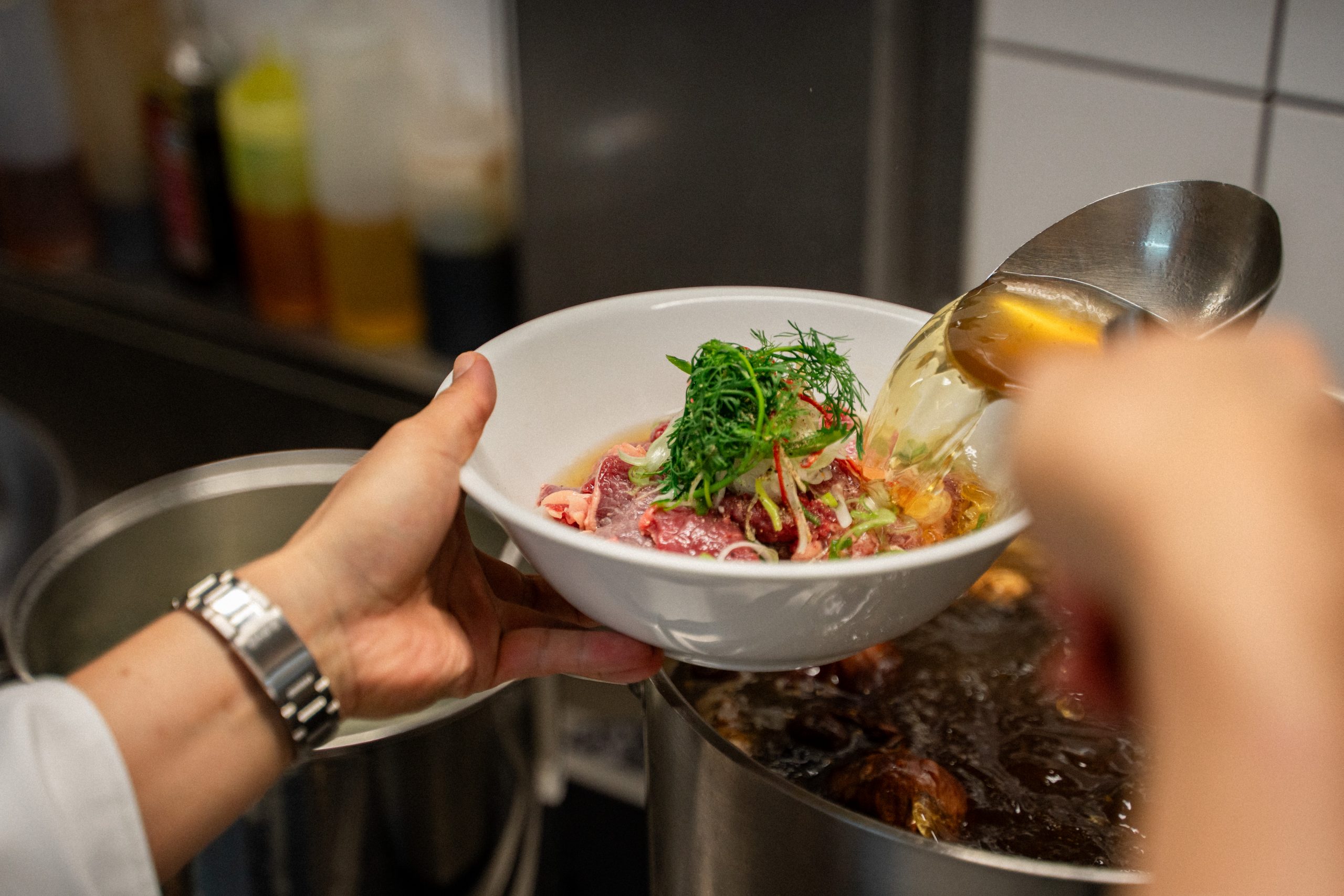 Hands hold a white bowl with sliced raw fish and fresh herbs as a ladle pours sauce into the bowl in a busy kitchen.