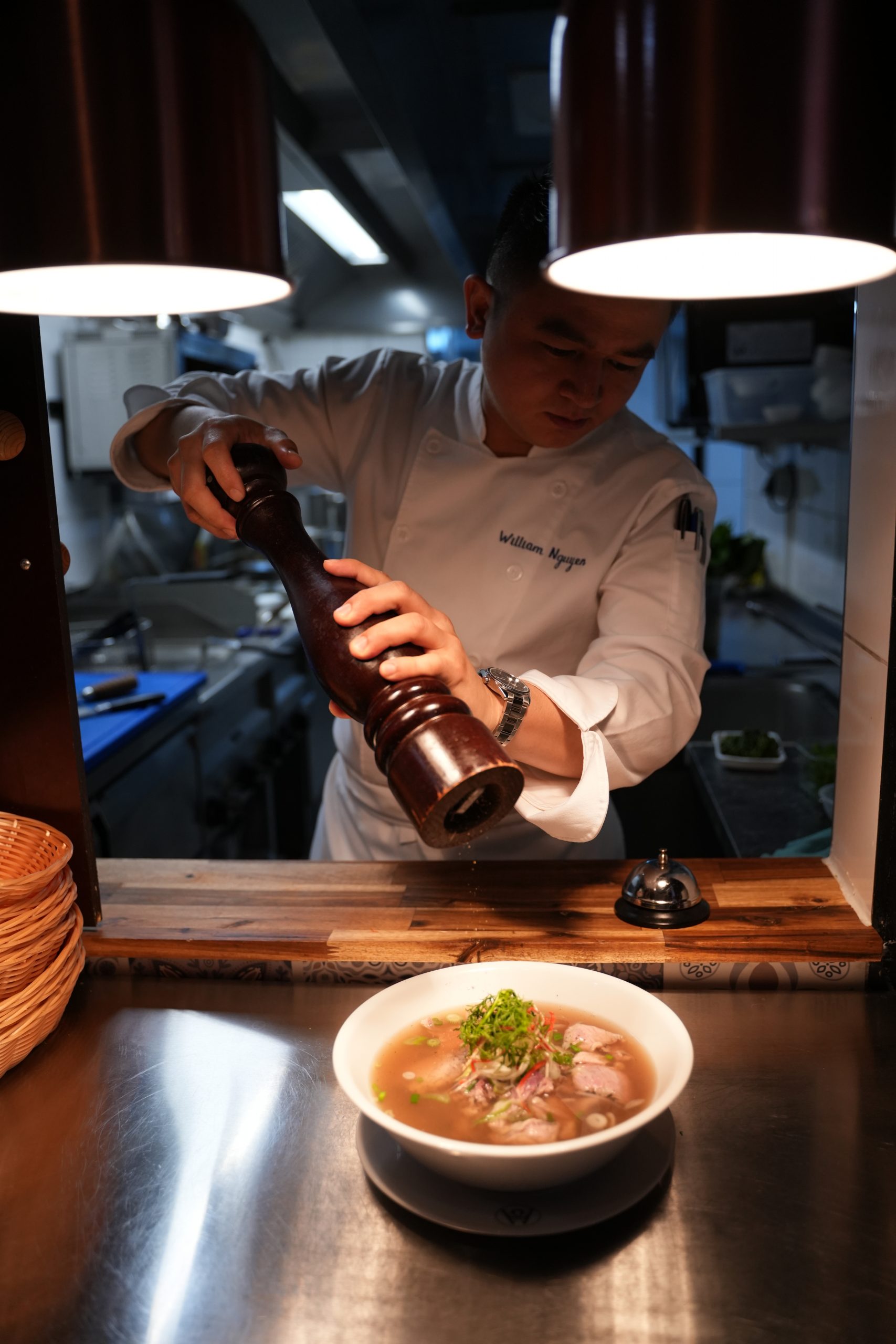 Chef in a white uniform seasons a bowl of soup at the pass, viewed through the kitchen window.