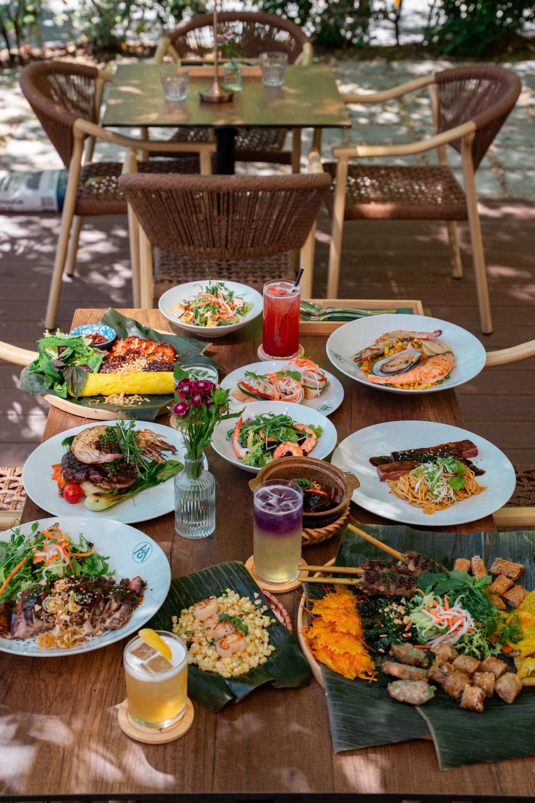 Outdoor wooden table full of diverse dishes and drinks, green salads, seafood, noodles, and skewers in a garden setting.