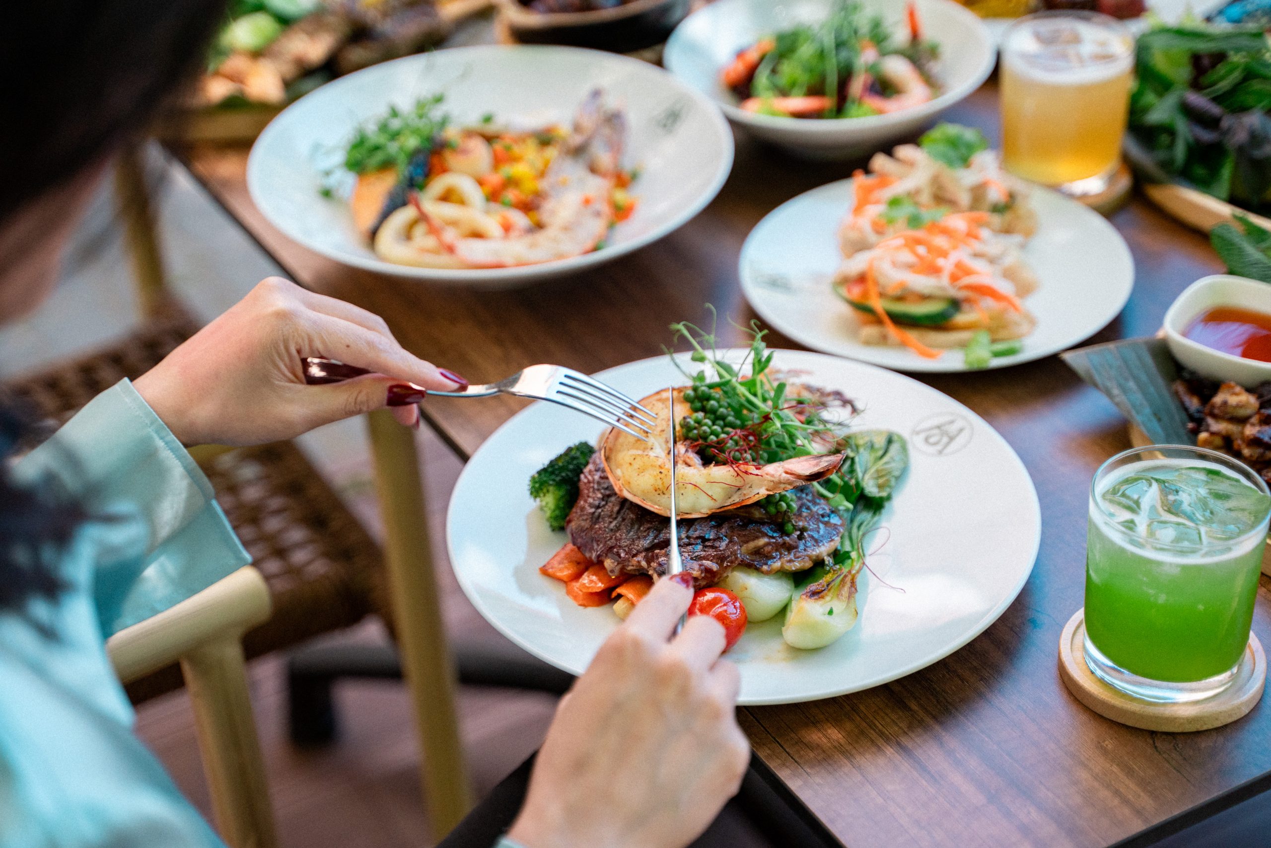 Person cutting into a plated steak topped with greens and seafood, with colorful side dishes on a wooden table and drinks nearby.